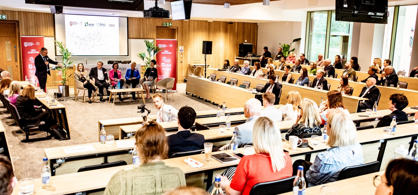 bright, modern lecture hall viewed from the back with presenter and seated panelists shown at the front and seated guests listening in
